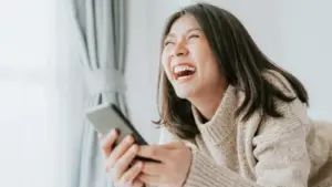 Wanita sedang melihat layar HP yang berisi kata-kata lucu dan wajahnya tertawa ngakak